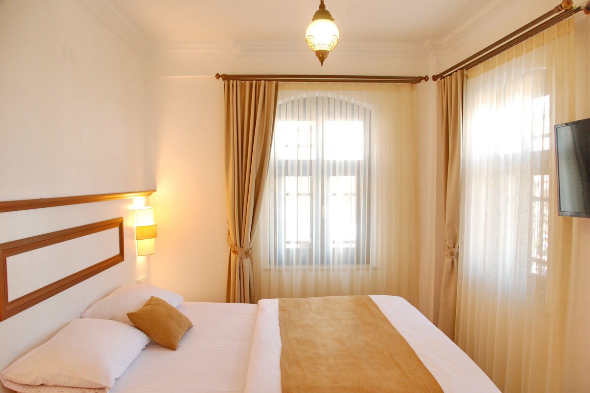 A hotel room with a bed, a headboard, a wall-mounted lamp, a window with curtains, and a television on the wall.