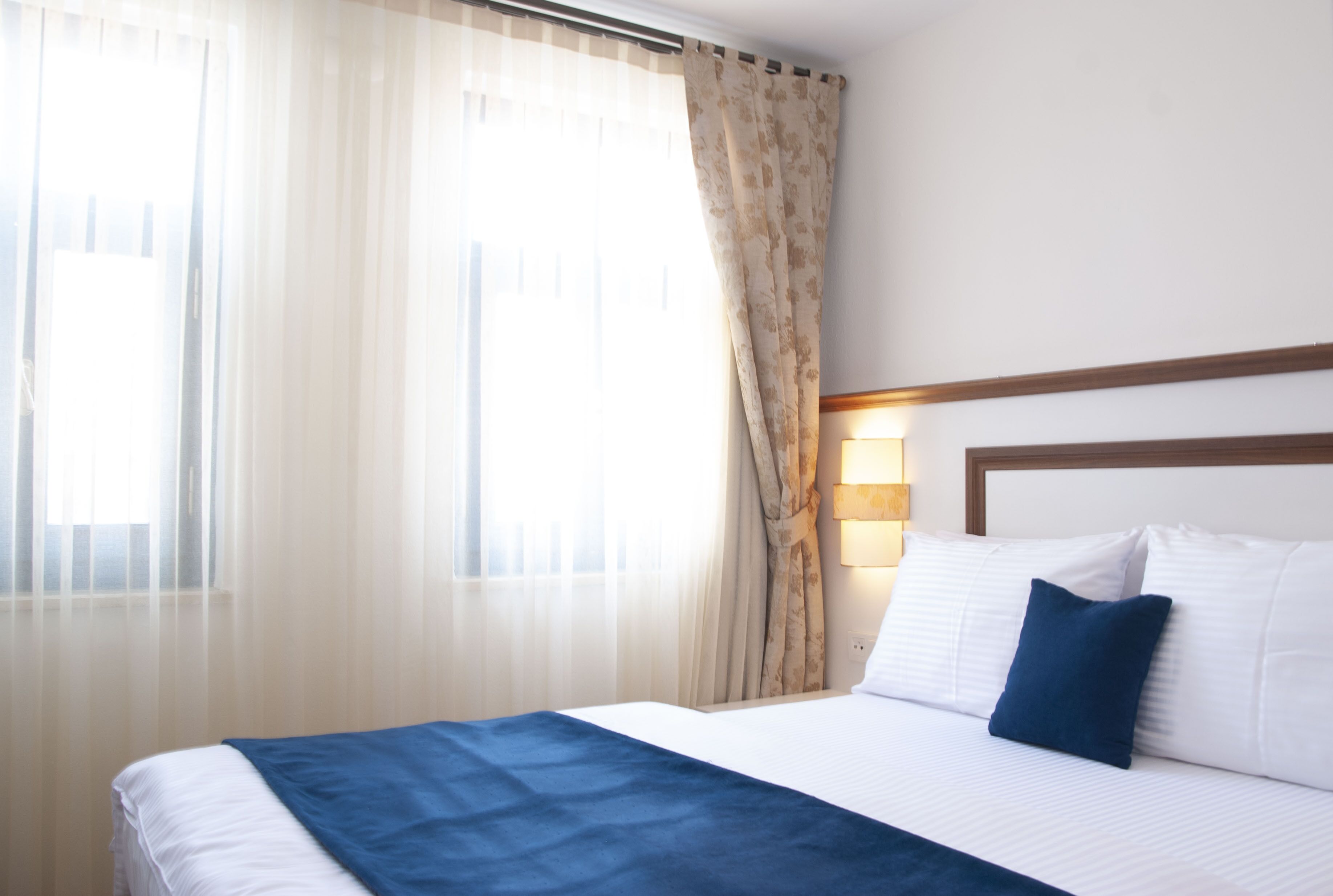 Habitación de hotel con cama, ventana con cortinas traslúcidas, lámpara de pared y cabecera de madera.