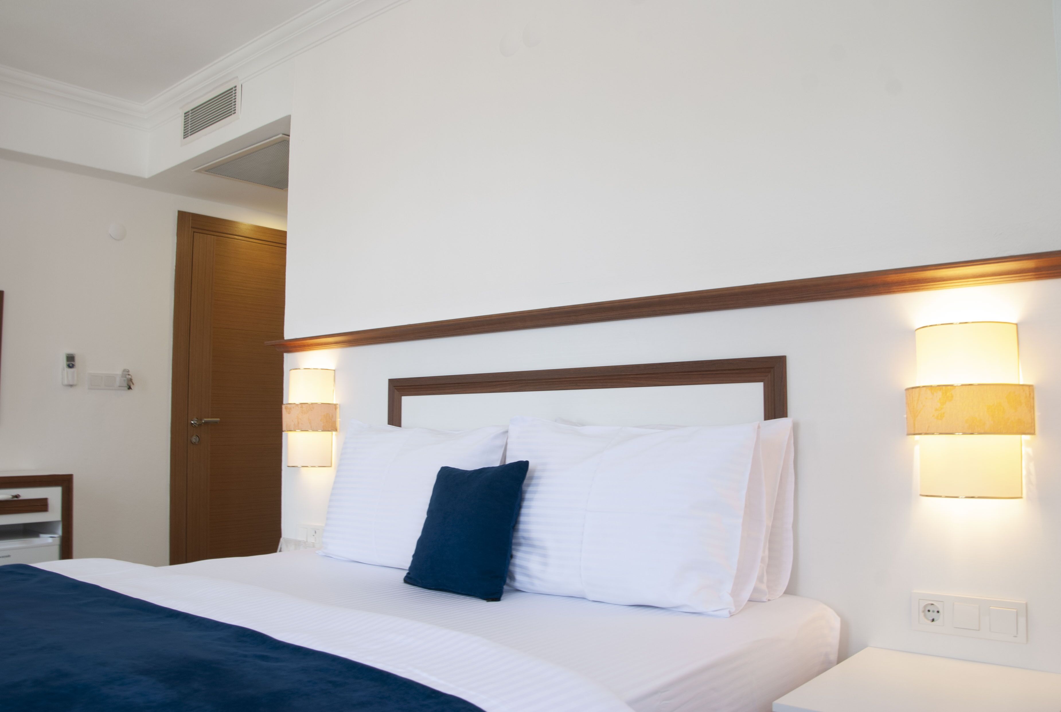 Una habitación de hotel con cama, lámparas de noche, cabecera de madera y una puerta.