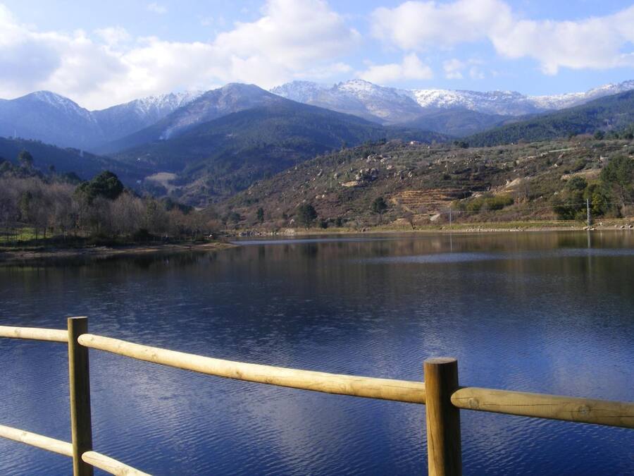 Un lago con montañas al fondo, una valla de madera en primer plano y un cielo despejado.
