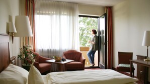 Habitación de hotel con cama, dos sillones, una mesa con un florero y vistas al exterior a través de una puerta.
