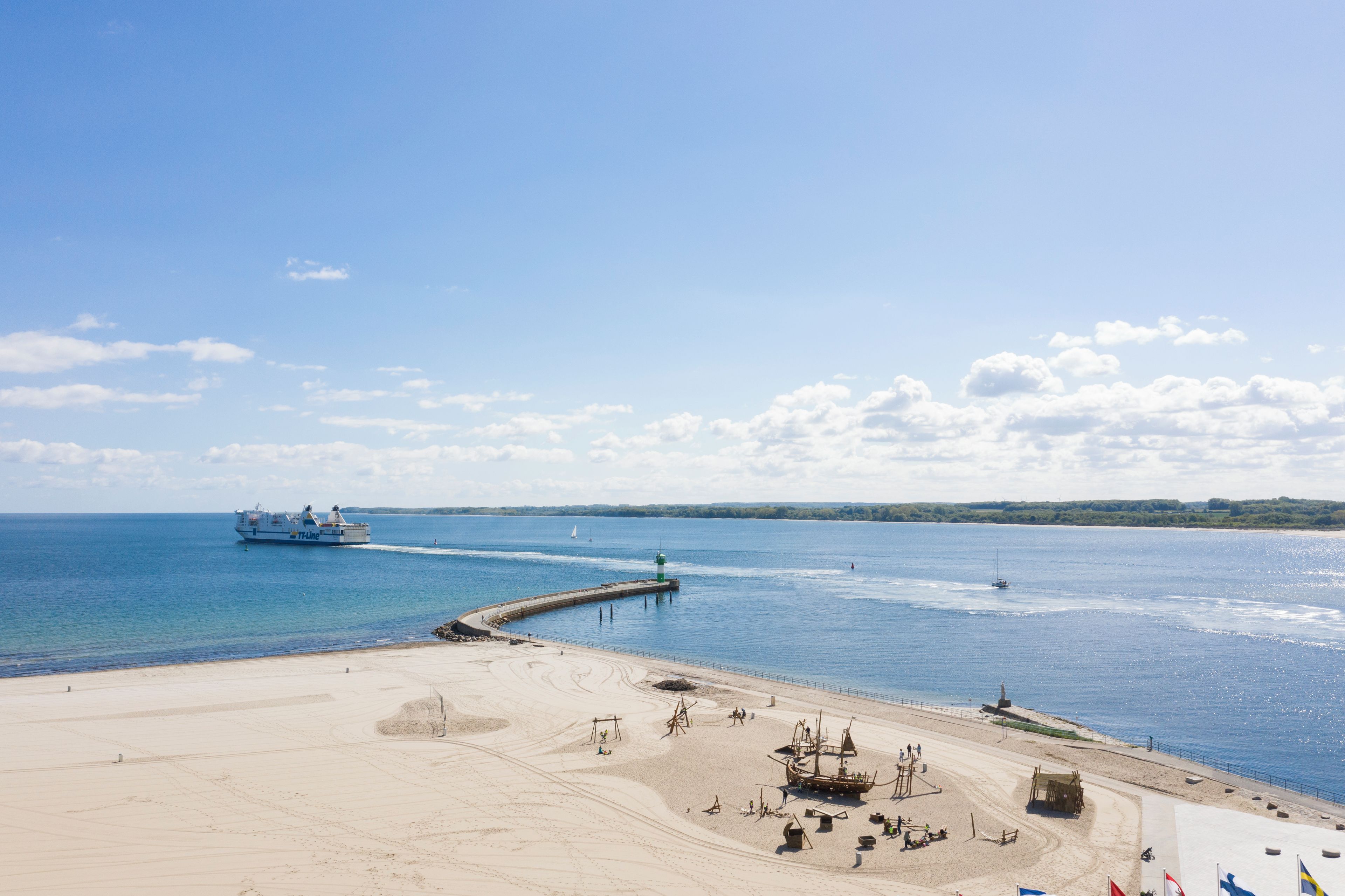 Uma praia com um píer que se estende para o mar, um navio grande passando e uma área arenosa com algumas construções.