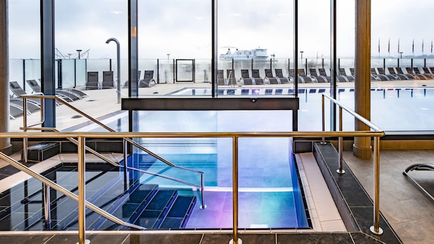 A modern indoor pool area with clear glass walls, a central pool, and sun loungers. - aja Travemuende (Lübeck)