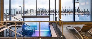 A modern indoor pool area with clear glass walls, a central pool, and sun loungers. - aja Travemuende (Lübeck)