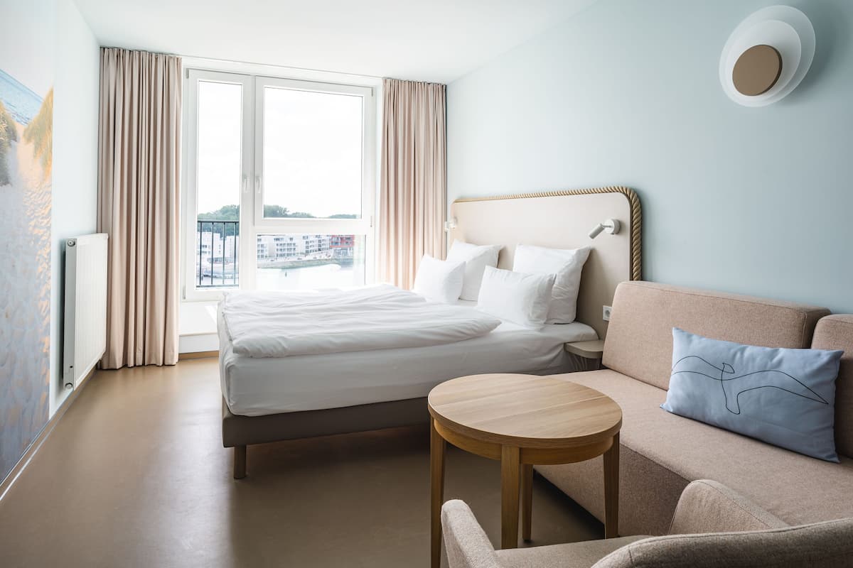 Une chambre d’hôtel avec un lit, un canapé, une petite table et une vue sur la ville grâce à de grandes fenêtres.