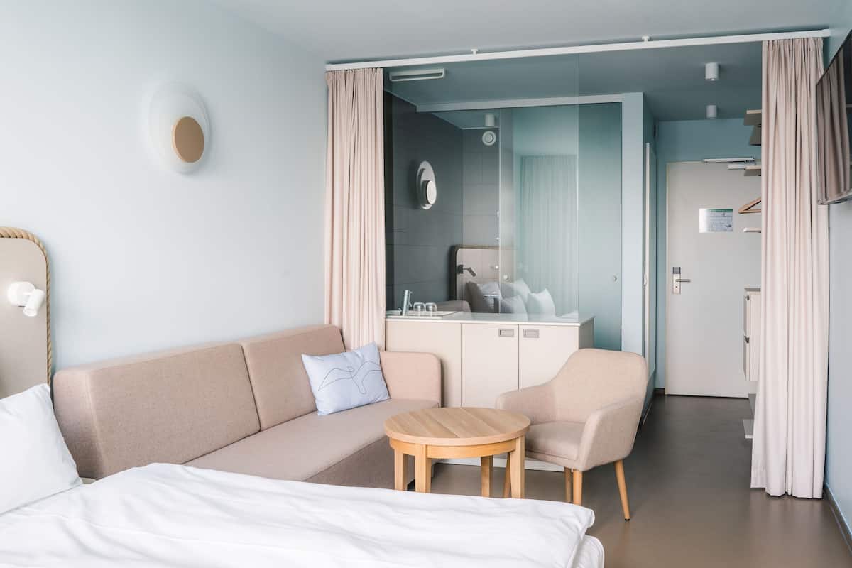 Une chambre d’hôtel moderne avec un lit, un canapé, une petite table en bois et, au fond, une kitchenette.