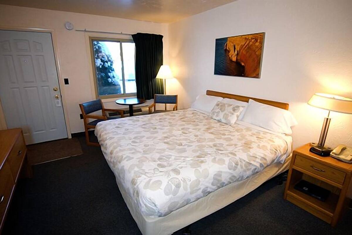 A hotel room with a bed, a desk, a chair, a small table, a lamp, and a painting on the wall.