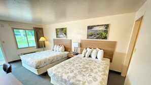 A hotel room with two beds, a desk, a lamp, and a window with a view of greenery. - Alger Falls Motel (Munising)