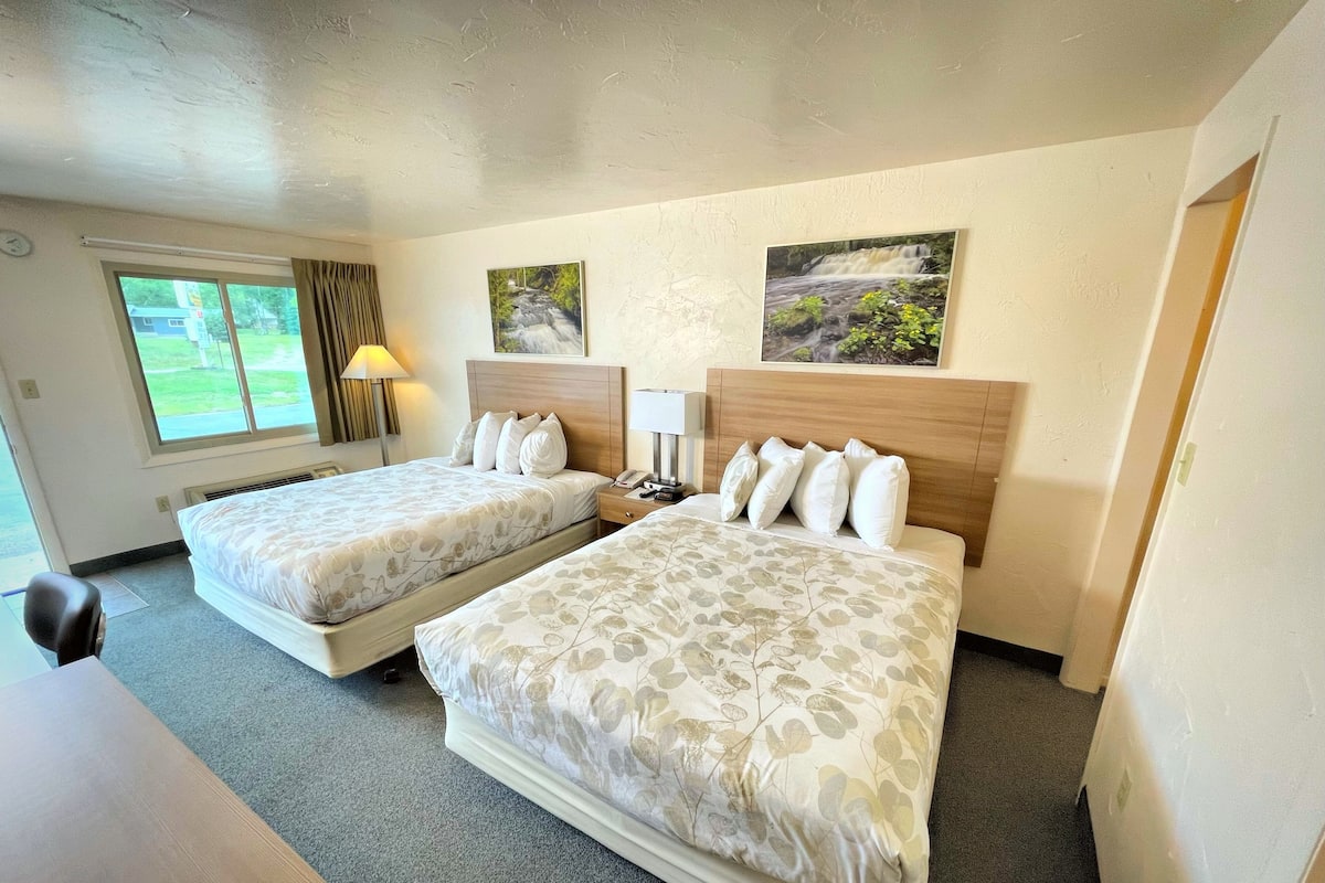 A hotel room with two beds, a desk, a lamp, and a window with a view of greenery.