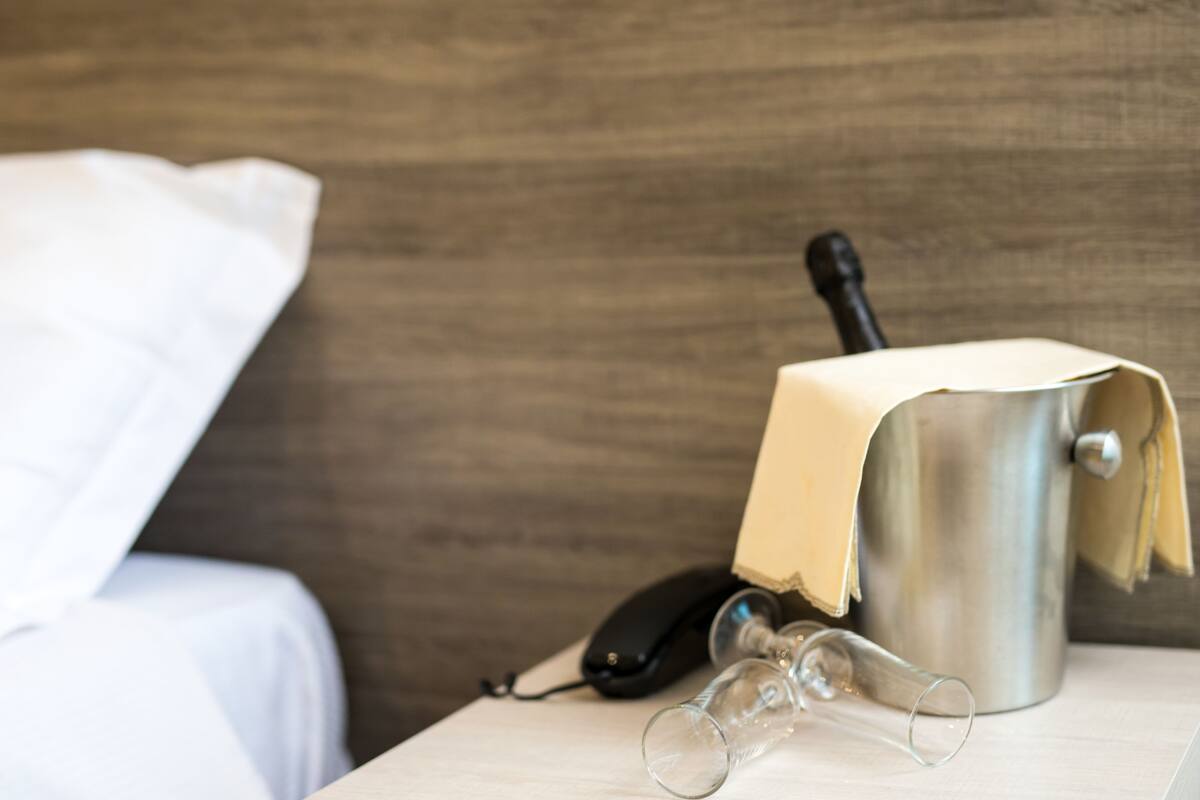 Habitación de hotel con cama, mesita de noche, teléfono y un recipiente metálico con funda de tela.