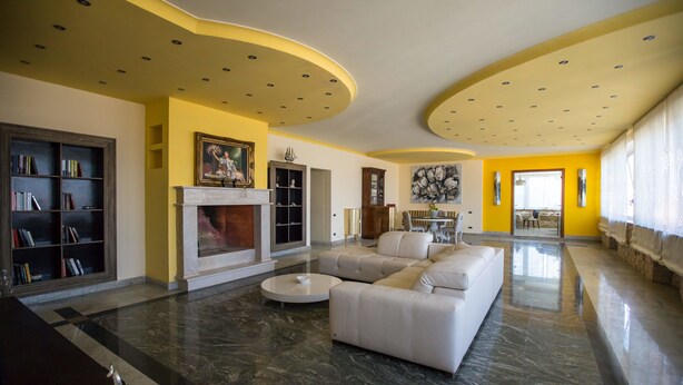 A spacious living room with a white sofa, a round coffee table, and a fireplace. There is a bookshelf and a dining area in the background. - Hotel Queen Daisy (Castellammare di Stabia)