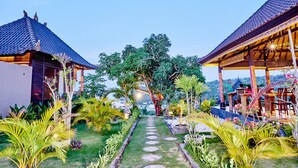 A pathway leading to two traditional-style buildings with thatched roofs, surrounded by lush tropical gardens and a clear sky. - Sunrise Huts Lembongan (Lembongan Island)