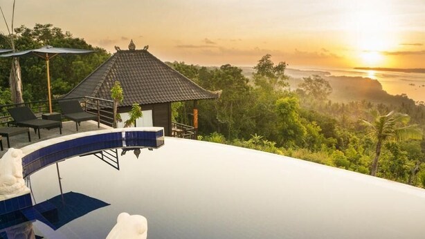 A infinity pool with a white statue, a small pavilion, and a view of the sunset over a forested landscape. - Sunrise Huts Lembongan (Lembongan Island)