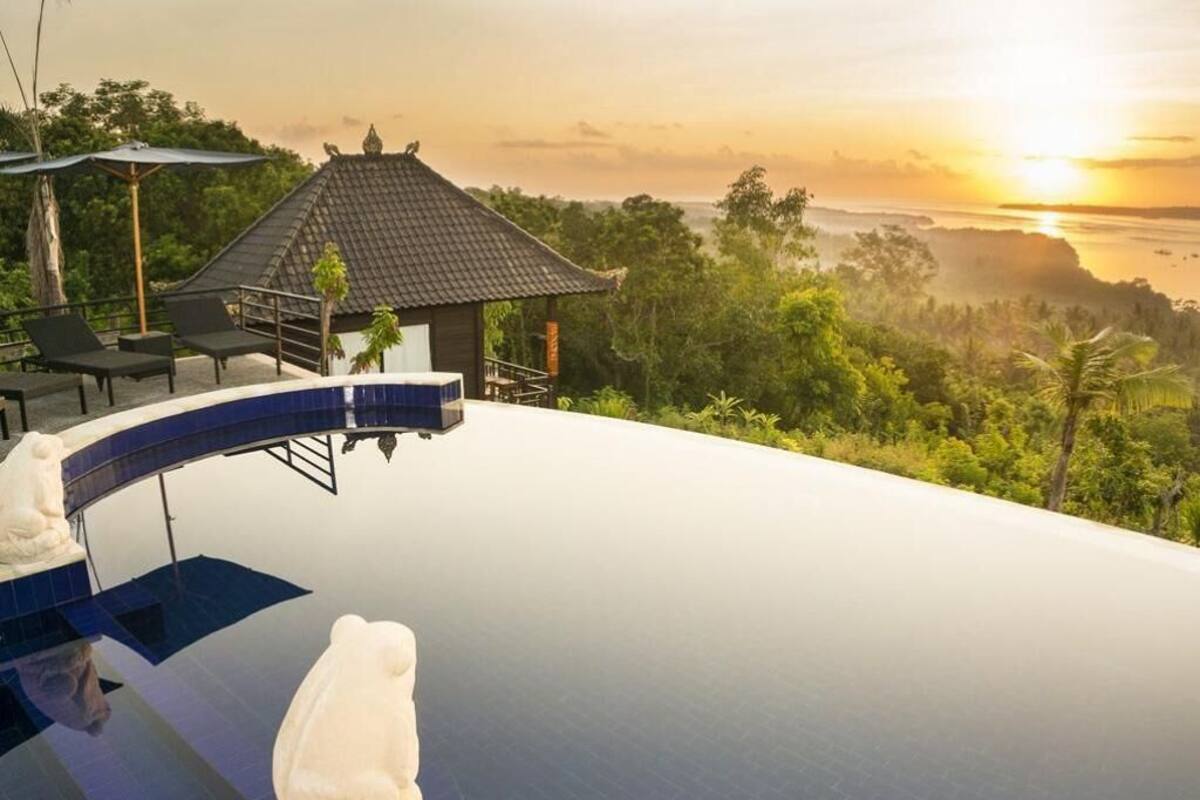 Ein Infinity-Pool mit einer weißen Statue, einem kleinen Pavillon und Blick auf den Sonnenuntergang über einer bewaldeten Landschaft.