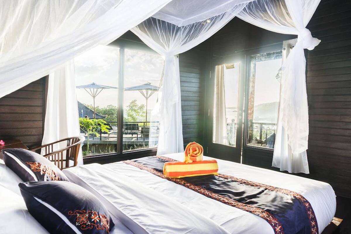 Ein Schlafzimmer mit Bett, weißen Vorhängen, einem Fenster und einem Balkon mit Gartenmöbeln.