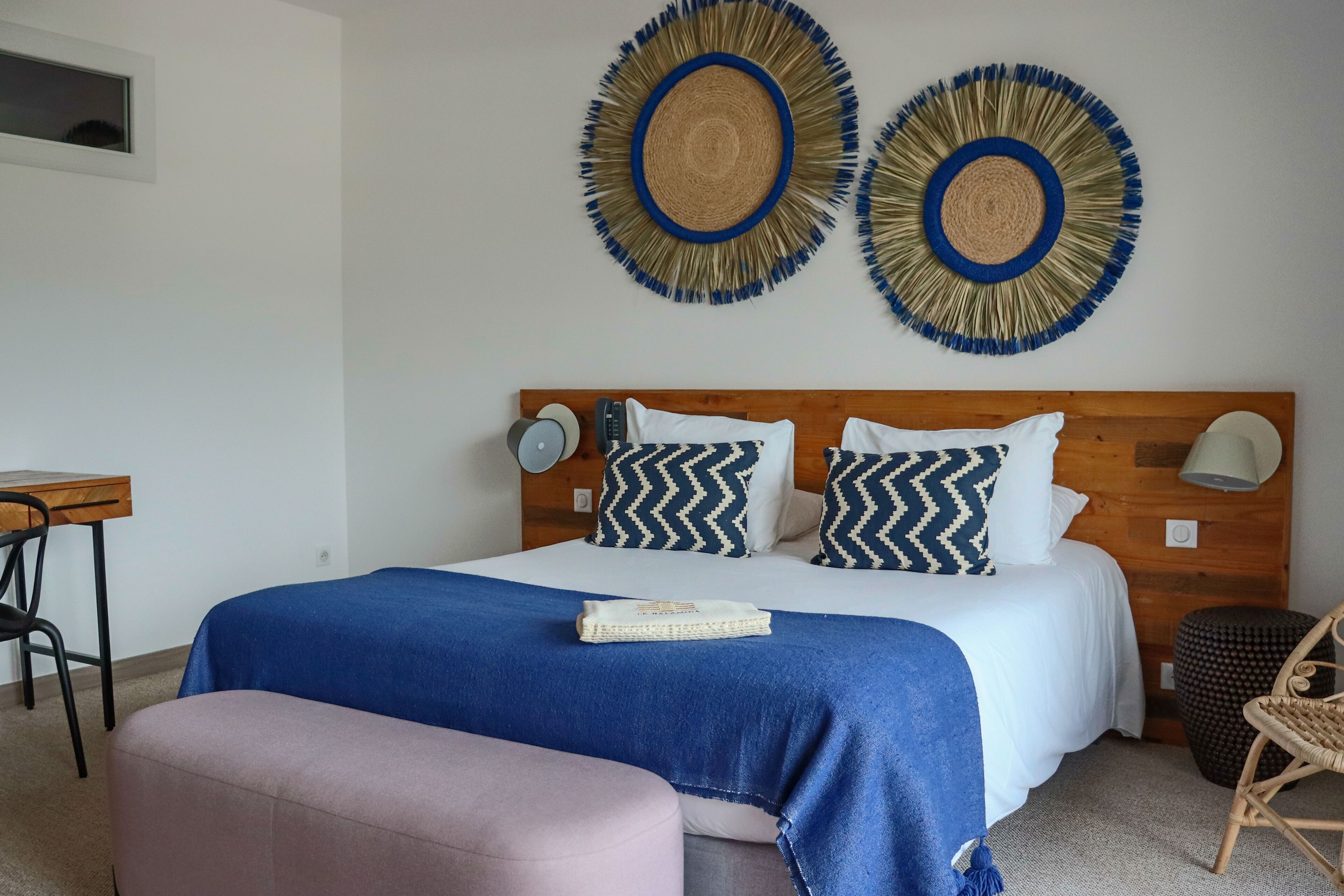 Une chambre à coucher avec une tête de lit en bois, deux décorations murales circulaires en fibres tressées, un lit avec une literie bleue et blanche, une table de chevet avec une lampe et une chaise.
