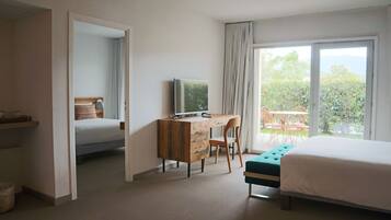 A hotel room with a bed, a desk with a computer, a chair, a window with curtains, and a view of greenery outside.