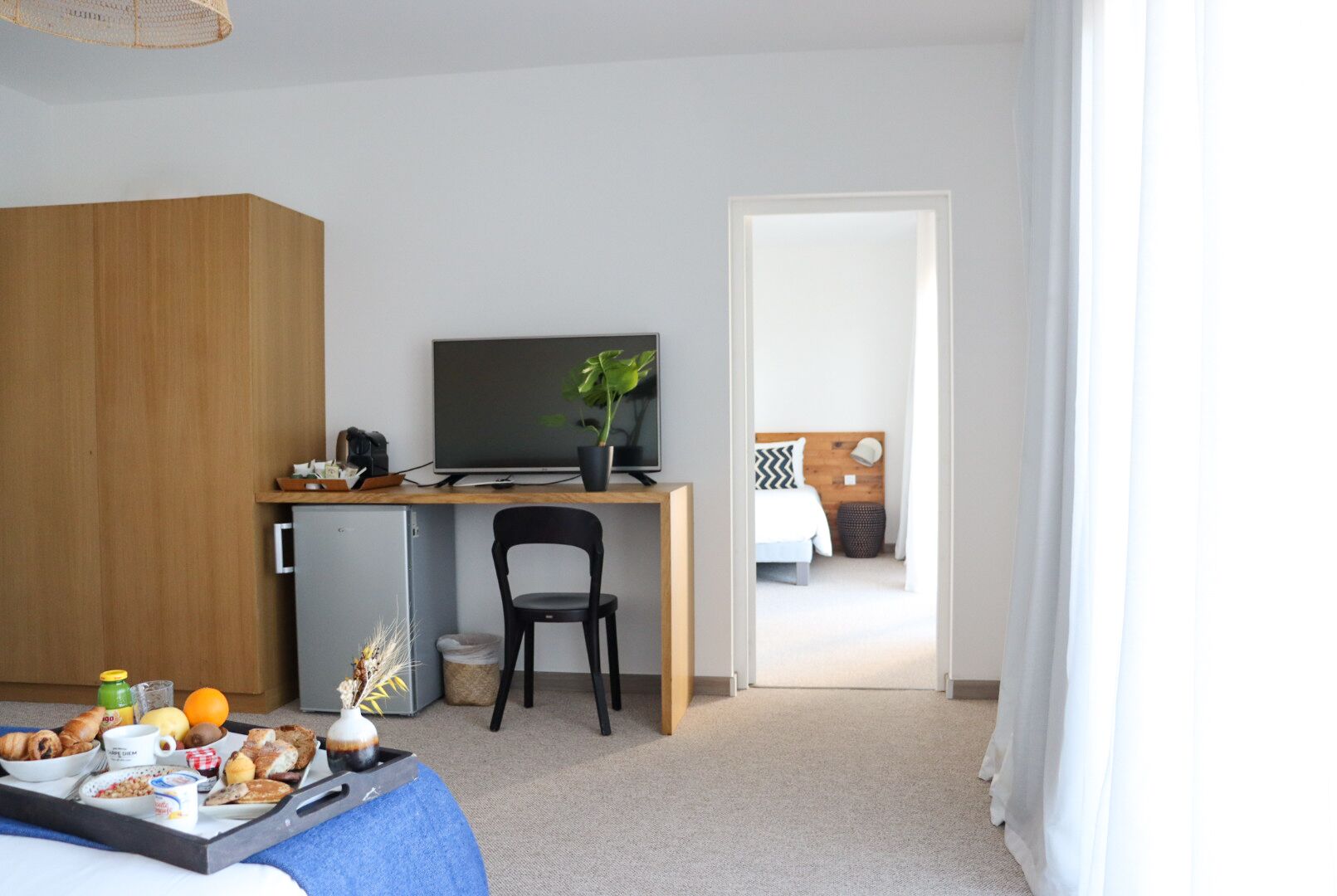 A hotel room with a wooden wardrobe, a desk with a computer and a plant, a bed with a headboard, and a breakfast tray on the floor.