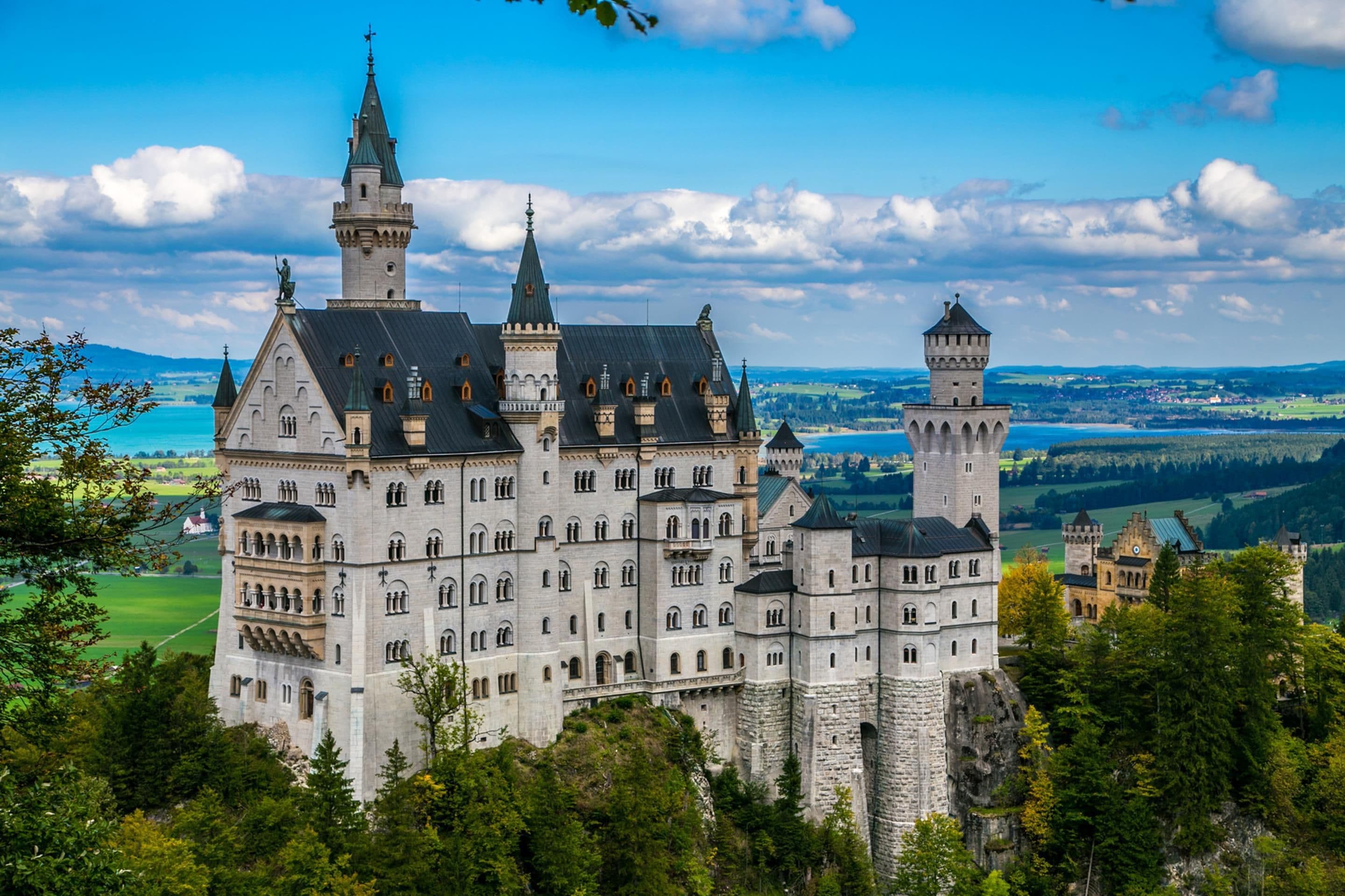 A large, historic castle with multiple towers and a prominent central building, surrounded by lush greenery and a scenic landscape.