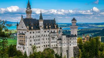 A large, historic castle with multiple towers and a prominent central building, surrounded by lush greenery and a scenic landscape.
