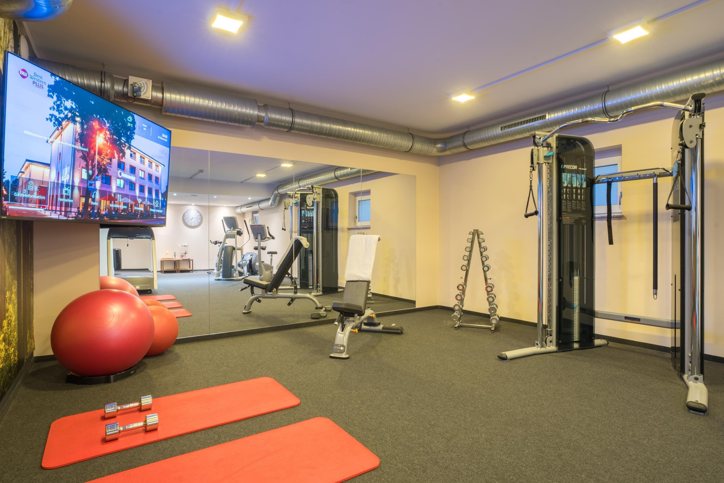 Un gimnasio con equipo de ejercicio, un televisor mostrando un hotel y un espejo que refleja la sala.