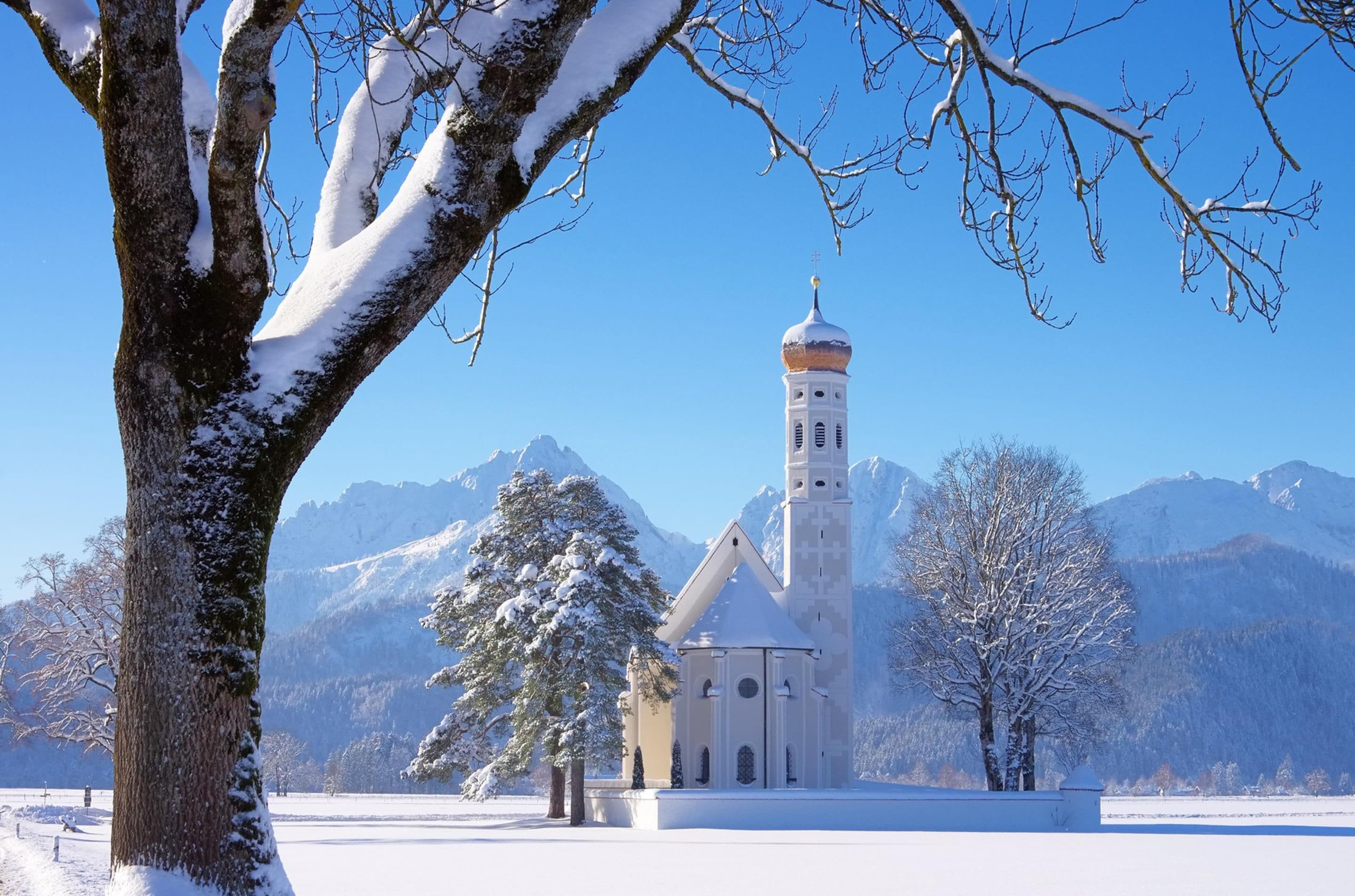 A church with a tall bell tower, surrounded by snow-covered trees and mountains in the background.