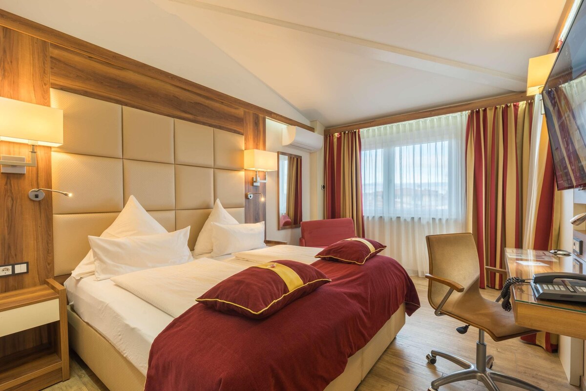 A hotel room with a large bed, a desk, a chair, and a window with curtains.