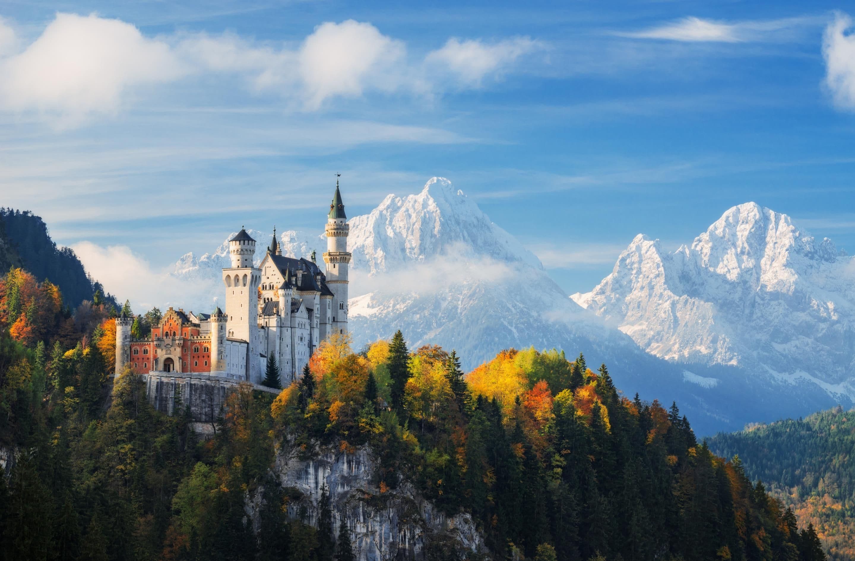 Un château entouré d’un feuillage automnal et de montagnes aux sommets enneigés.