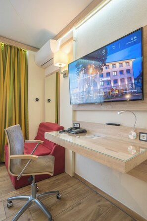 Une chambre d’hôtel avec un téléviseur fixé au mur, diffusant une vue sur la ville, un bureau avec une chaise et un canapé rouge.