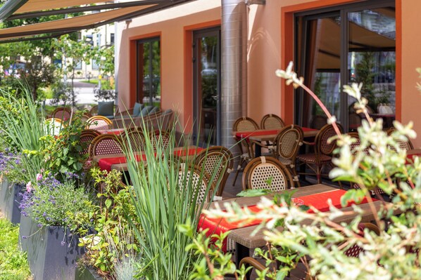 Un café en plein air avec des meubles en osier, des coussins rouges et des plantes en pot.
