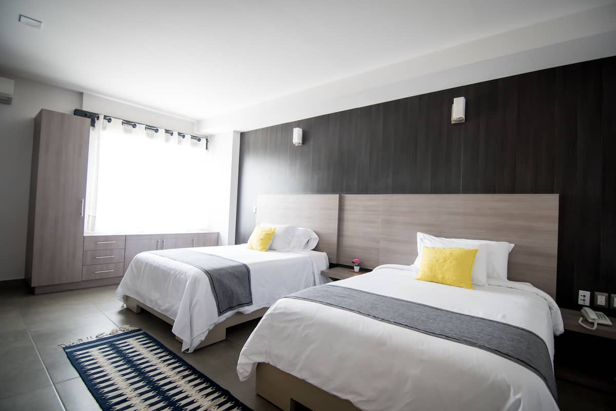 A hotel room with two beds, a large window with curtains, a dark wood headboard, and a bedside table with a phone.