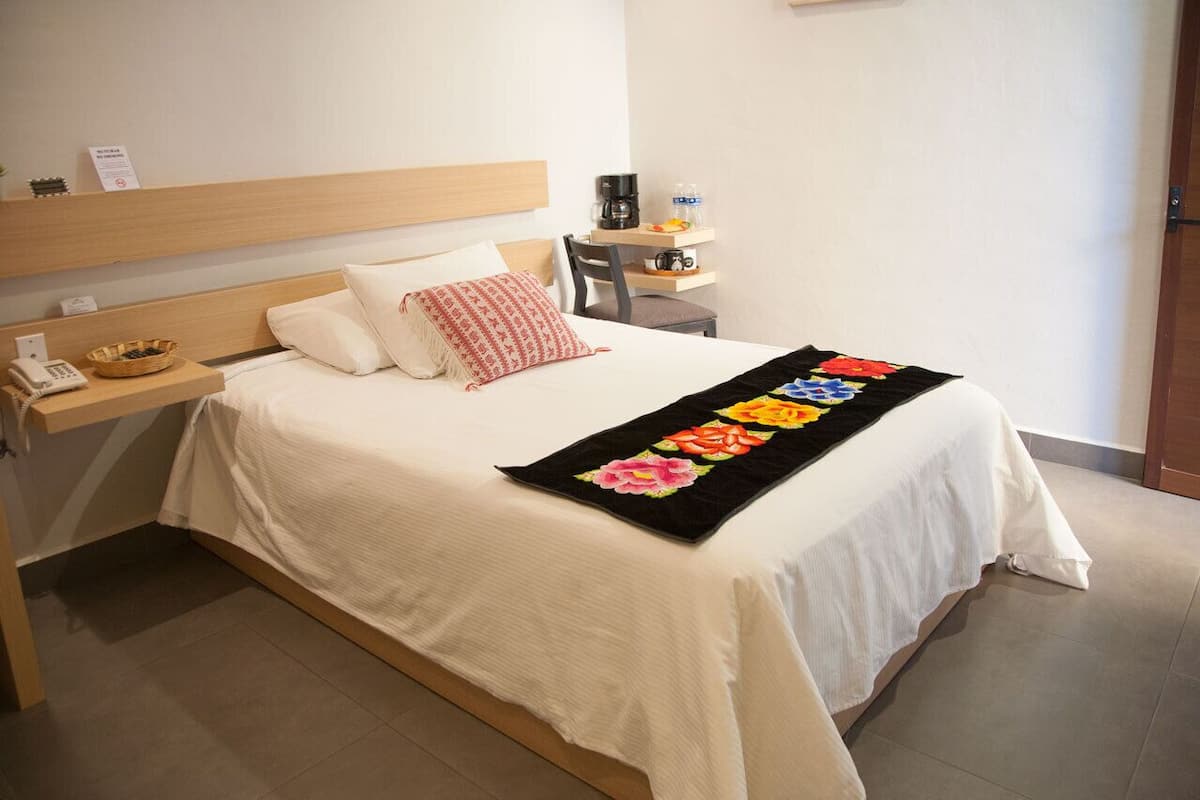A neatly made bed with a floral bedspread, a bedside table with a basket and a phone, and a small shelf with a coffee maker and a water bottle.