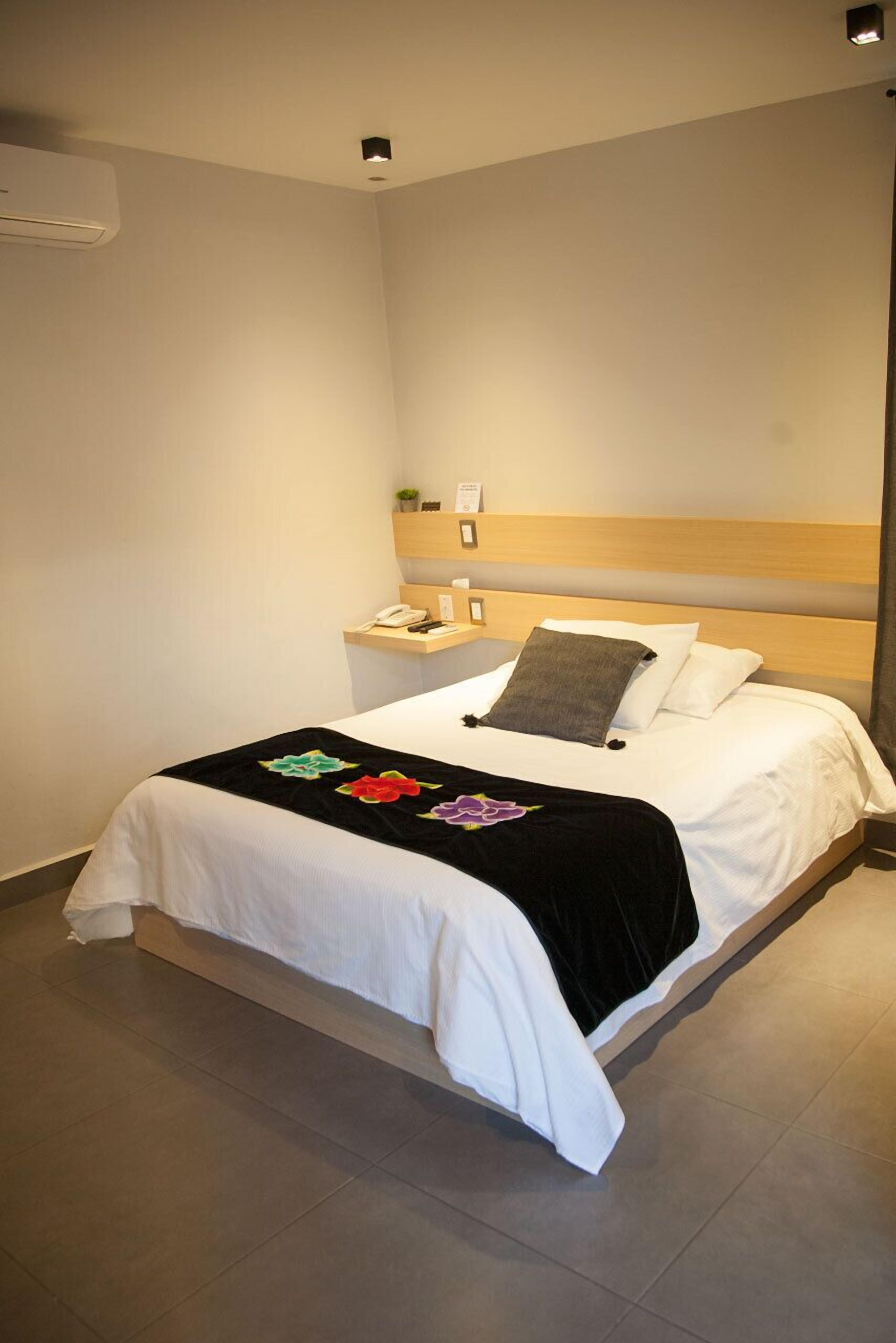 Una cama bien hecha con colcha blanca y negra, cabecera de madera y mesita de noche con lámpara y teléfono.