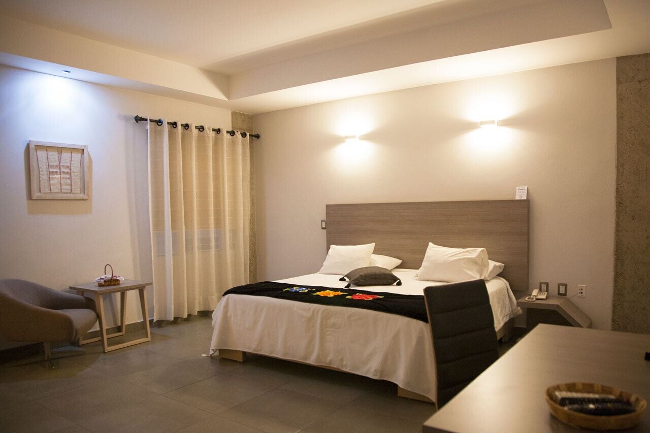 A hotel room with a bed, bedside table, chair, and a small table with a basket.