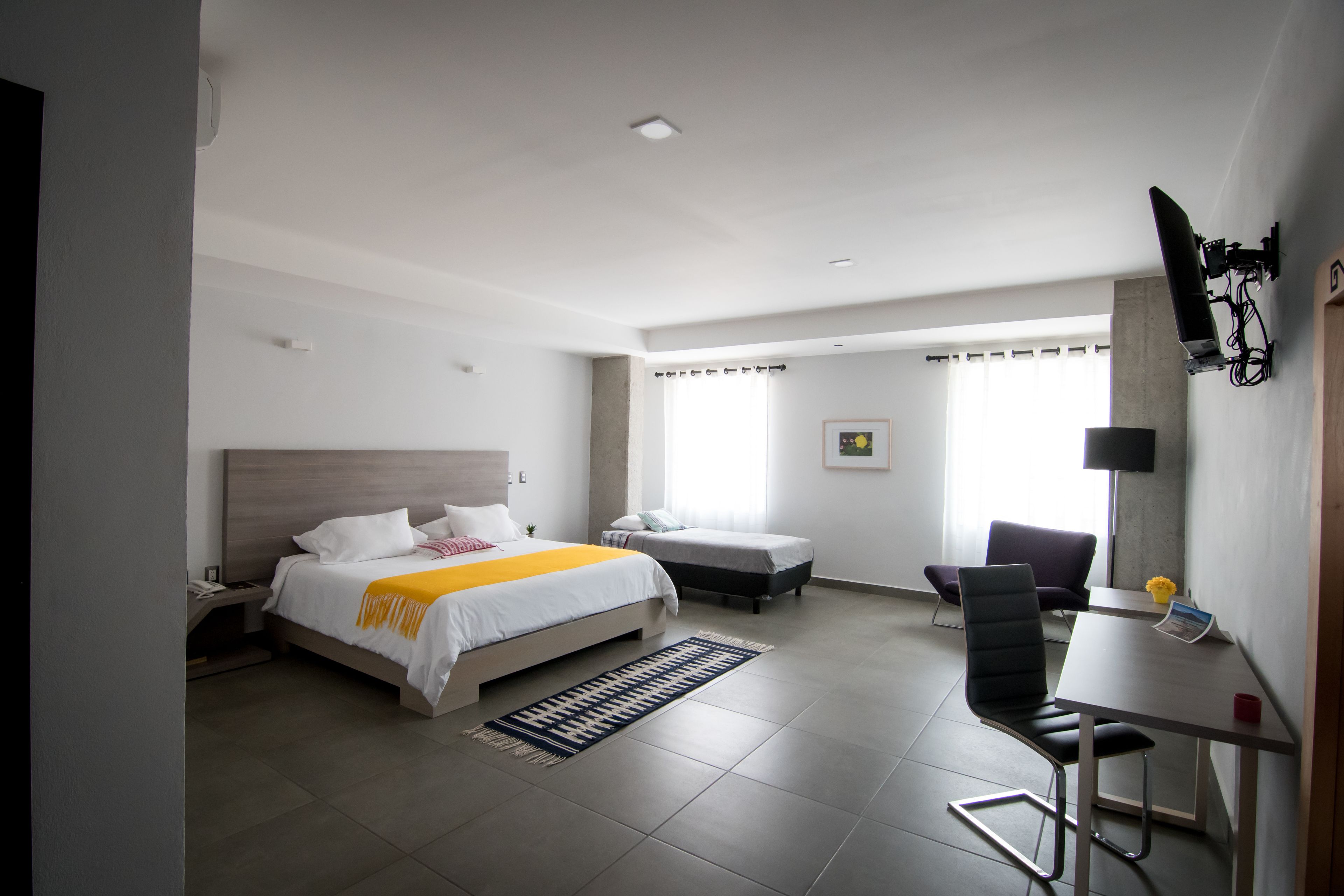 A hotel room with a bed, a desk, a chair, a small table, a lamp, and a window.