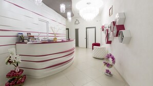Un área de recepción con un mostrador blanco curvo, detalles en rosa y un candelabro. Hay plantas en macetas y una silla blanca al fondo.