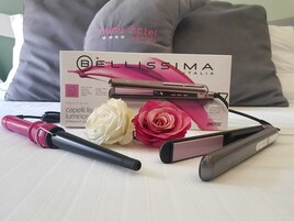 A hair straightener, a box labeled 'Bellissima Italia', and two artificial roses on a bed.