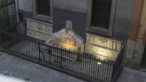 Un banco de piedra con intrincados grabados, una pequeña fuente y una valla metálica frente a un edificio con dos ventanas y una puerta.