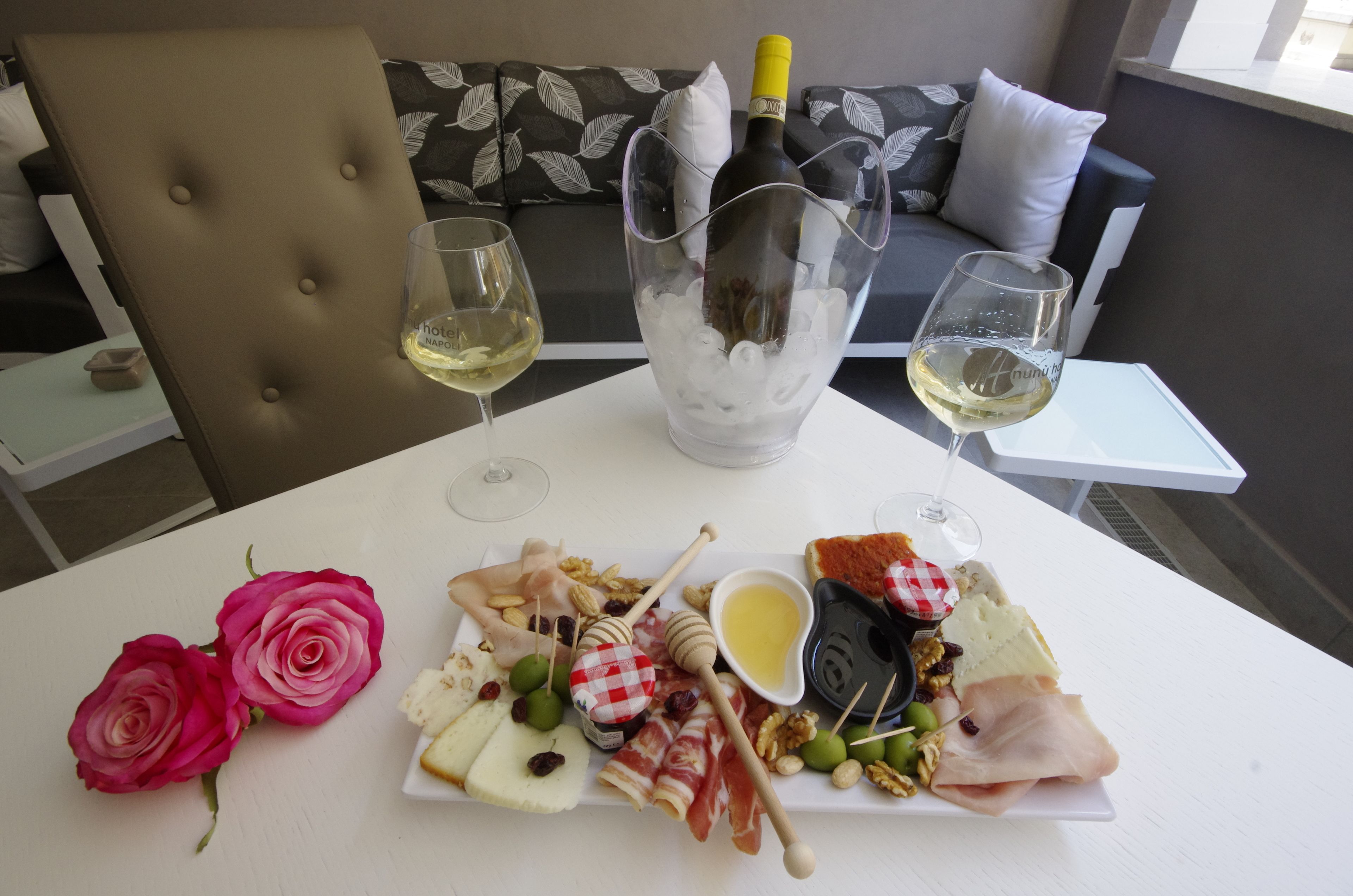 Un tavolo con una bottiglia di vino, due bicchieri di vino bianco, un piatto di formaggi, frutta e noci, e due rose.