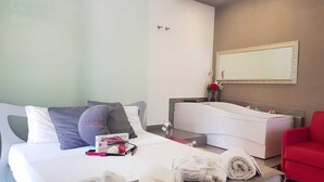 Habitación de hotel con una cama, una silla roja, un tocador blanco con espejo y un jarrón rojo con flores.