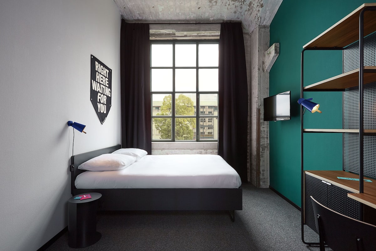 Habitación de hotel con cama, escritorio, silla, ventana con cortinas, televisor montado en la pared y un cartel en la pared.