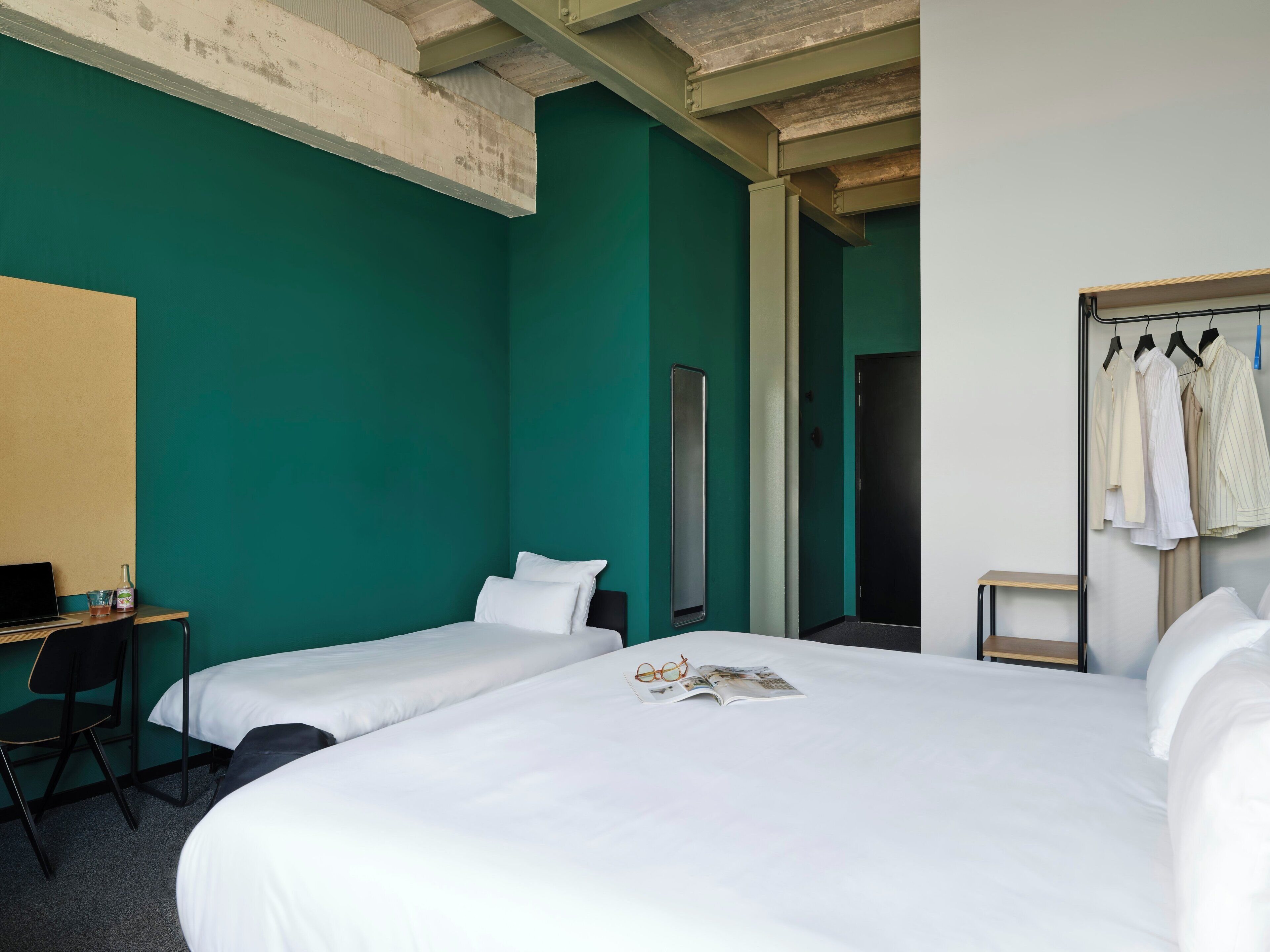 Habitación de hotel con dos camas individuales, un escritorio con silla, un armario con ropa colgada y una puerta que da a otra habitación.