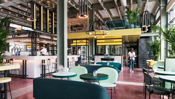 Zona de bar moderna con mostrador, taburetes y una sección de vinos y cócteles. Hay mesas y sillas para sentarse, y el espacio tiene techos altos con vigas a la vista e iluminación colgante.