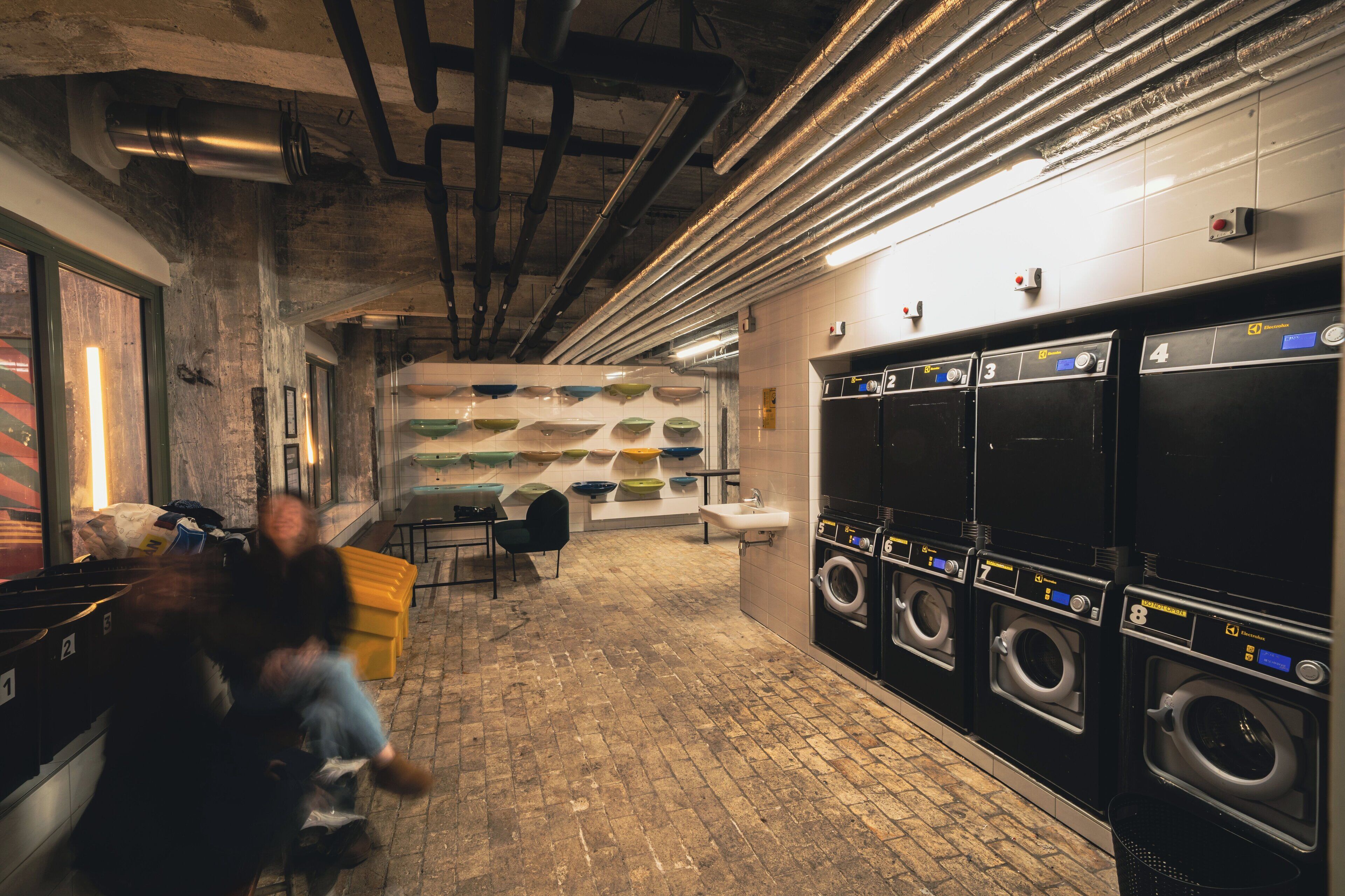 A public laundromat with multiple washing machines and dryers, a tiled floor, and a brick wall.