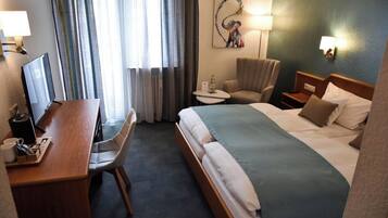 A hotel room with a bed, a desk with a chair, a small table, and a lamp.
