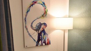 A hotel room with a beige armchair, a small white table with a lamp, and a painting of an elephant on the wall.