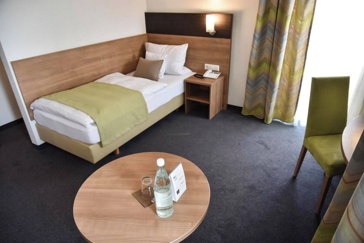 A hotel room with a bed, a bedside table with a lamp, a chair, and a round table with a bottle and glass.