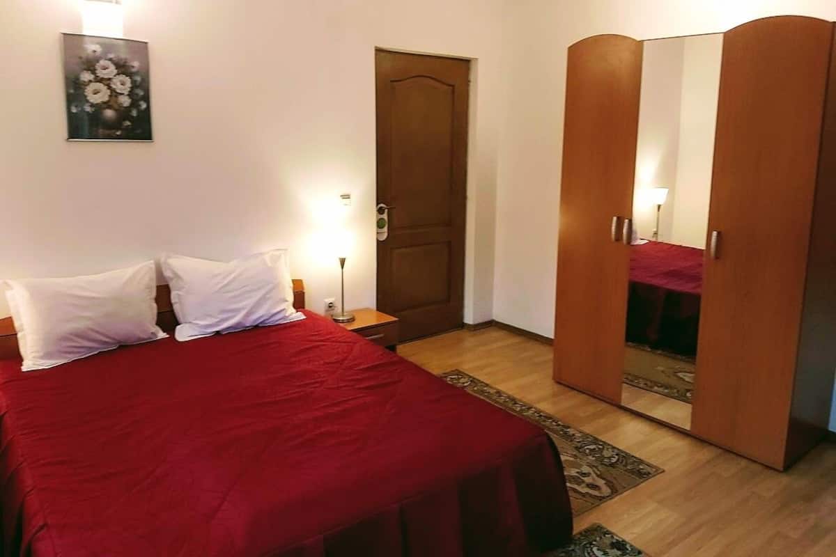 A bedroom with a bed, two bedside tables, a wooden wardrobe, a door, and a wall-mounted light fixture.