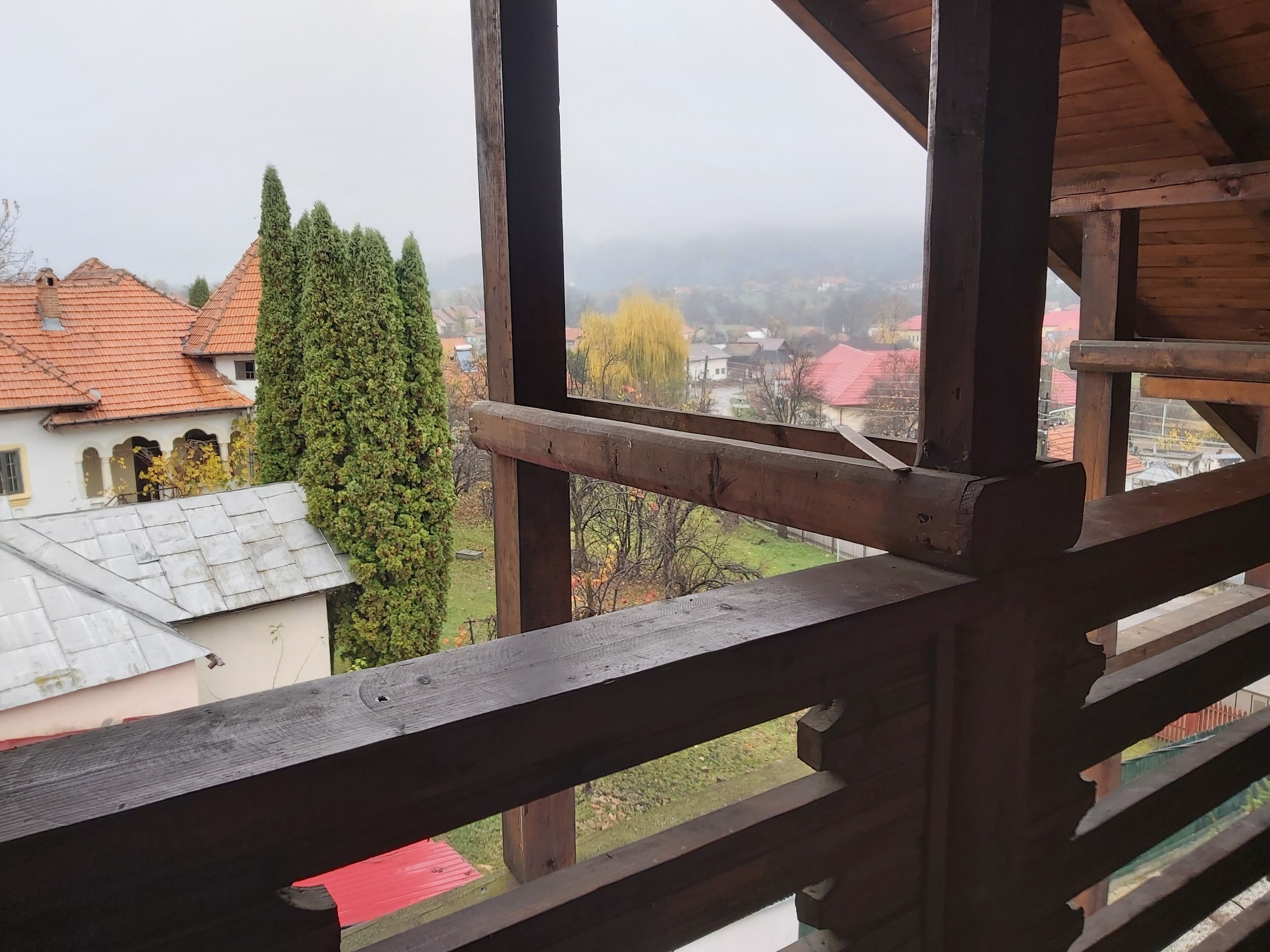 Vista desde un balcón de madera con barandilla, que da a una zona residencial con casas y un árbol alto.