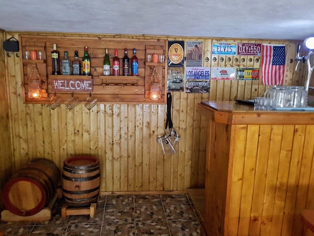 A bar area with wooden walls, a welcome sign, and various liquor bottles. There are wooden barrels and a tiled floor.